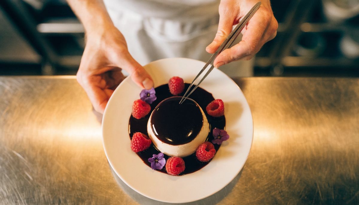 An overhead shot of the chef's hands arranging raspberries and edible flowers around the panna cotta with tweezers