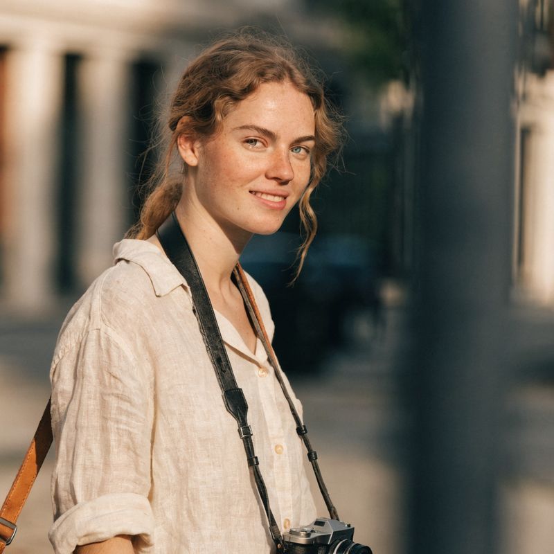 A female traveler in her late twenties with light brown shoulder-length hair, a cream linen shirt, and a small black camera on a leather strap