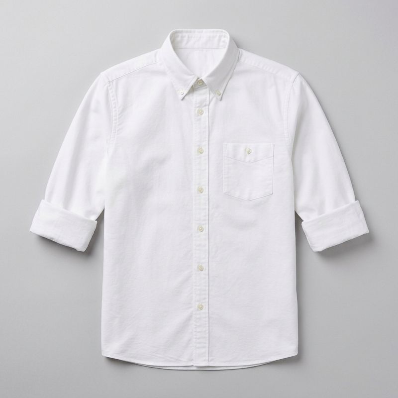 Flat-lay product photograph of a crisp white oxford button-down shirt with sleeves rolled to the elbows, on a plain pale grey background