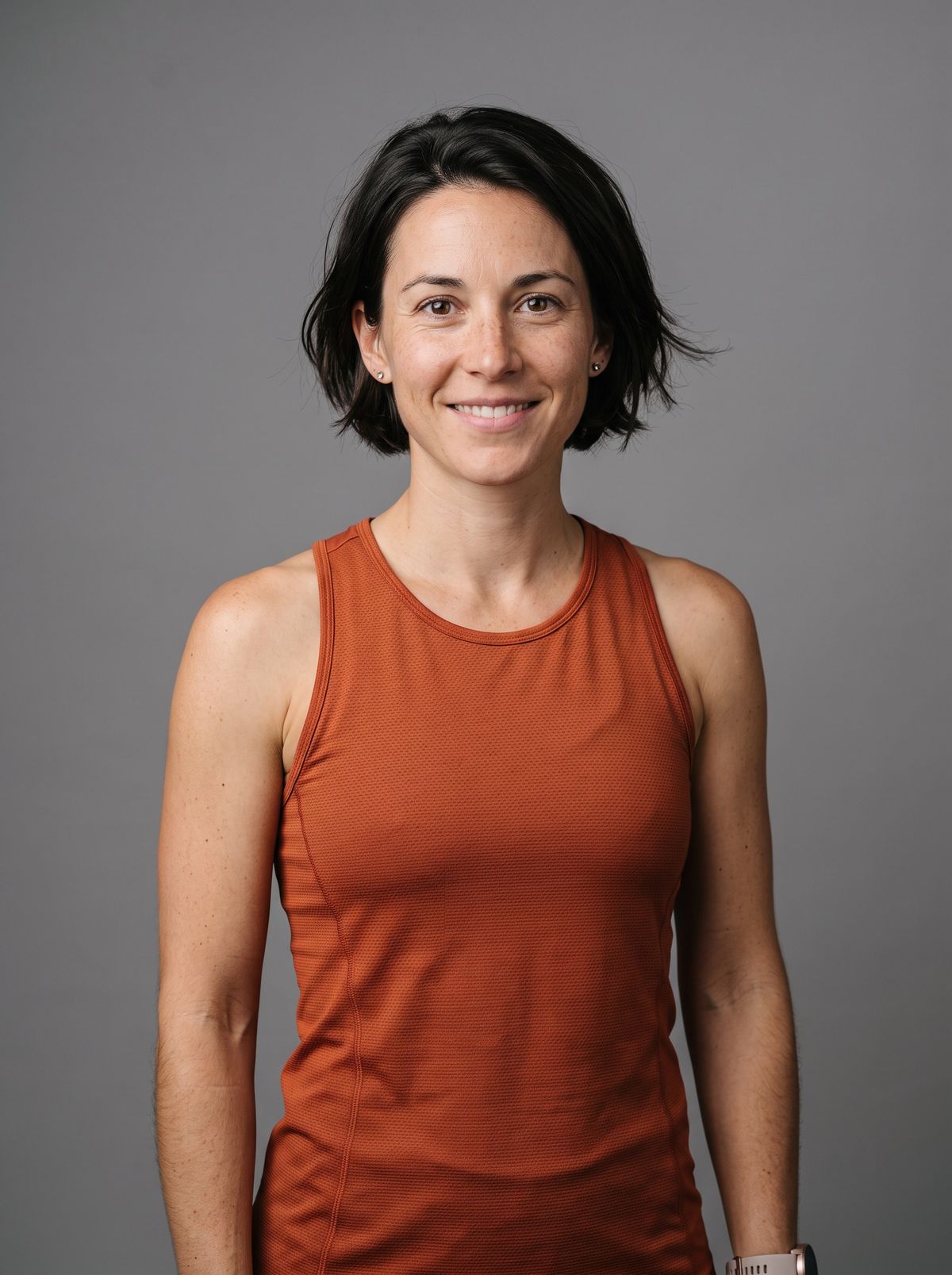 A studio portrait of a freckled trail runner with a short dark bob in a rust-orange running top against a gray backdrop