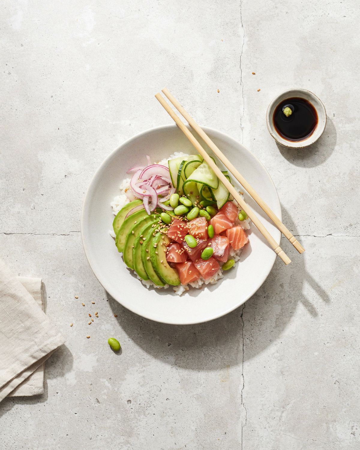 An overhead salmon poke bowl with rice, salmon, avocado, edamame and cucumber on a pale concrete table