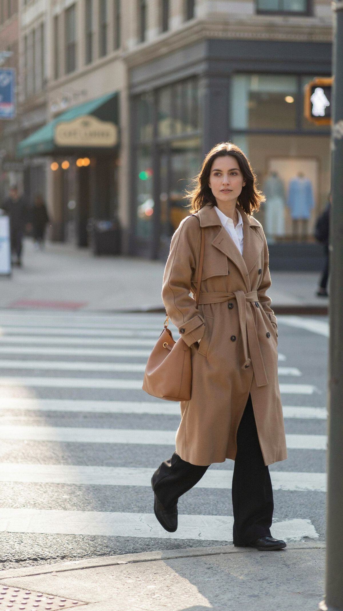 A woman in a camel trench coat and wide-leg black trousers crossing a sunlit crosswalk mid-stride, full figure in a vertical frame