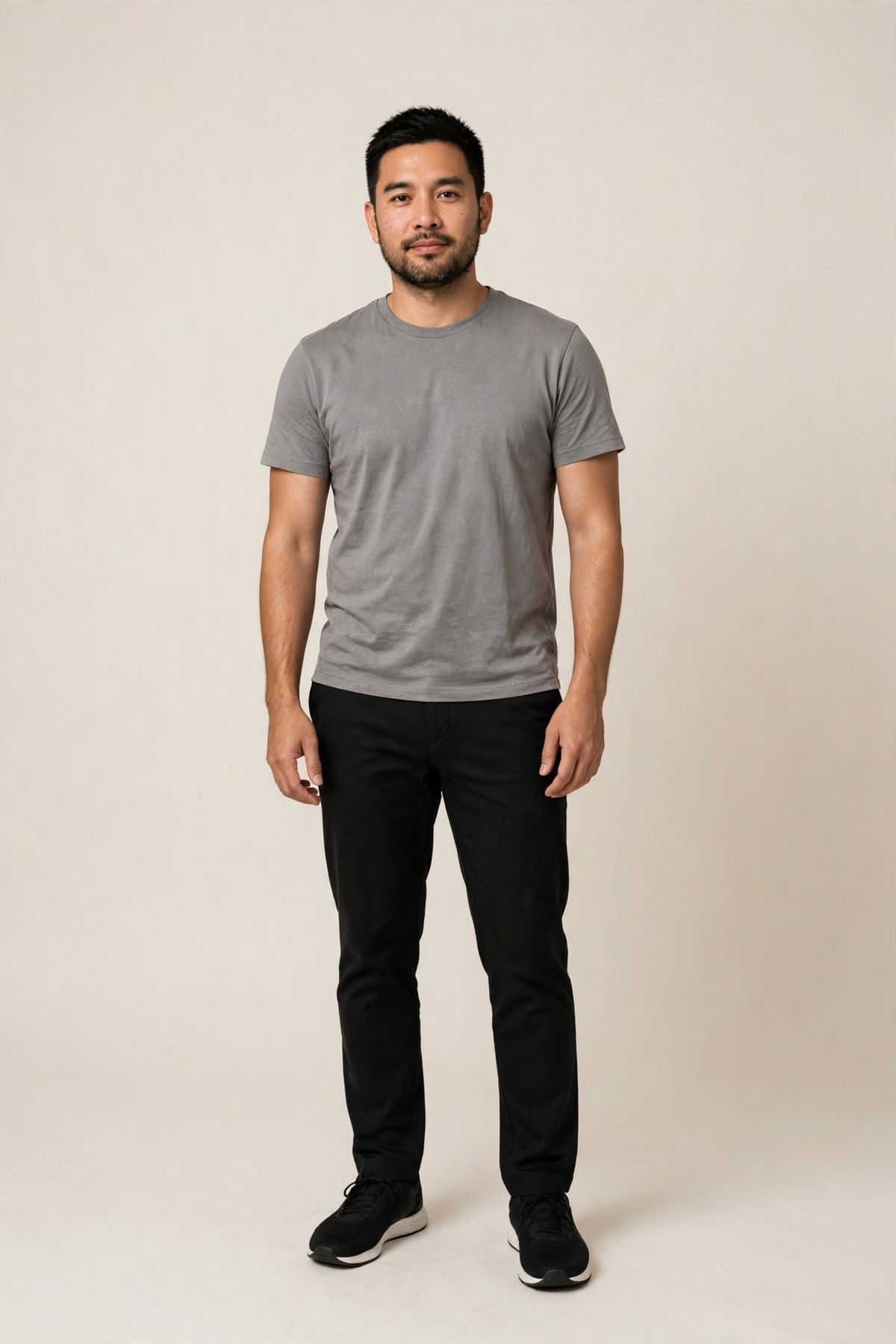 A full-length studio photo of a man in a grey crew-neck t-shirt and black trousers on a warm-white backdrop