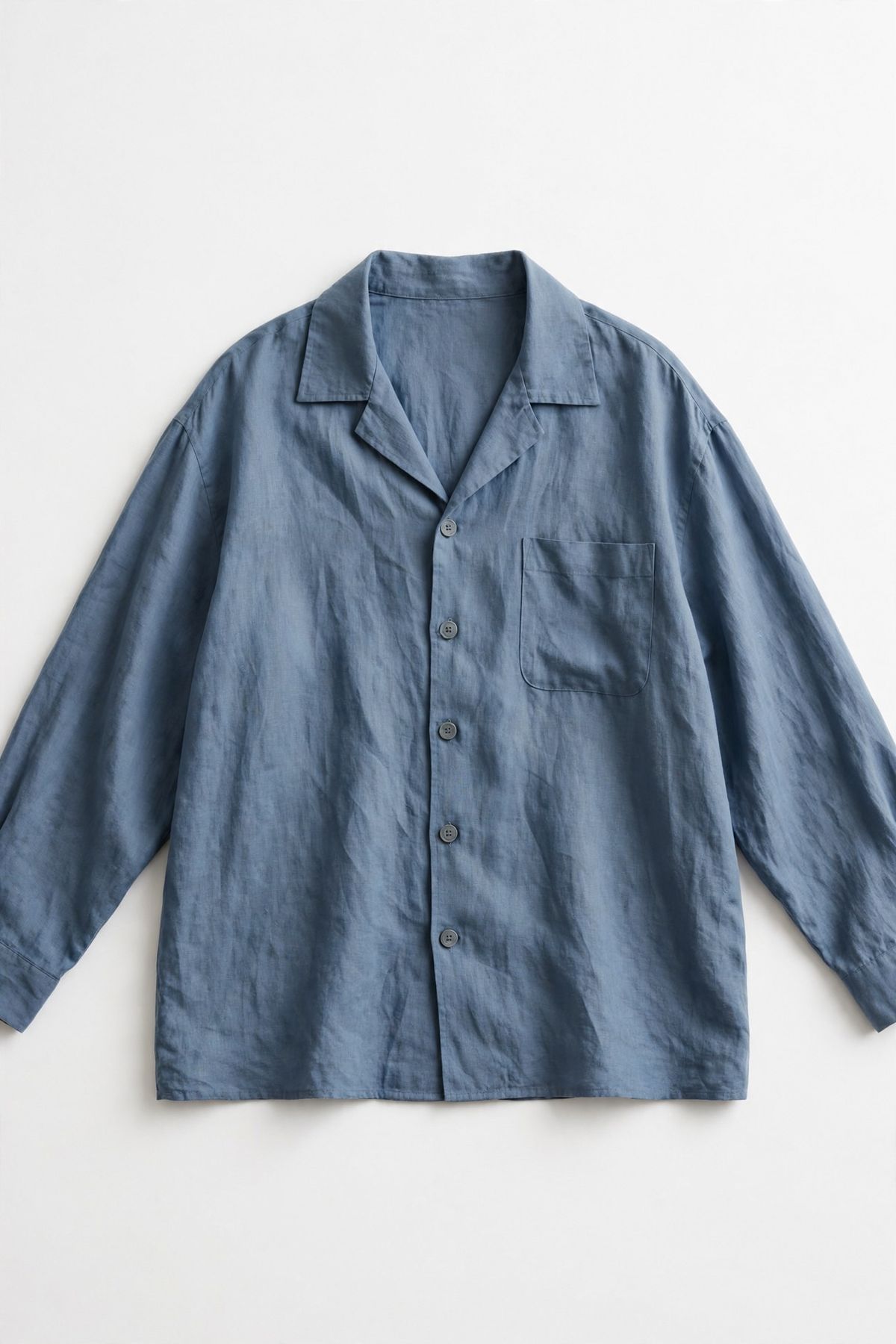 A vertical flat-lay of a dusty blue linen overshirt with a chest pocket and matte grey buttons on a white background