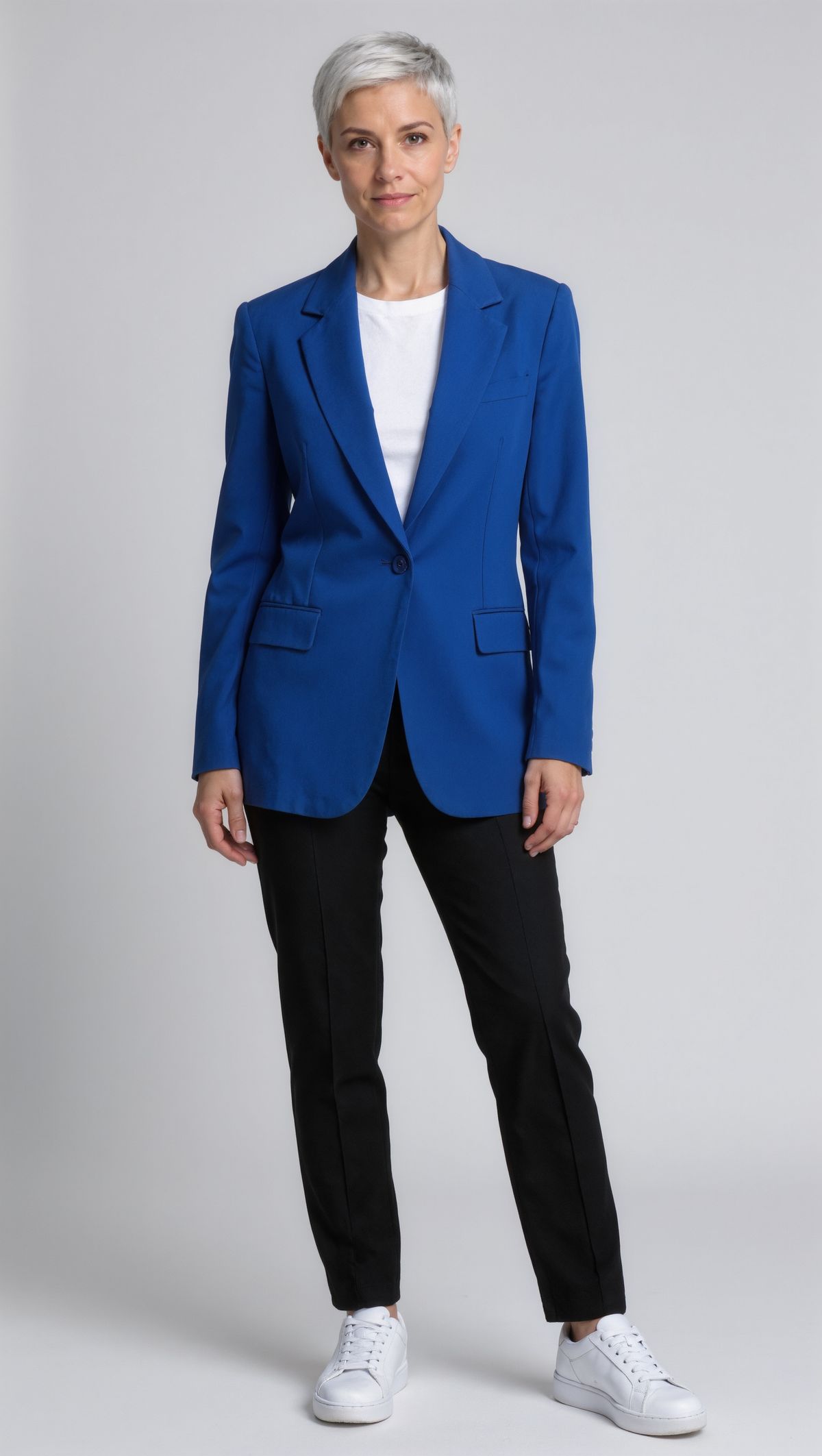 Front-facing full-length portrait of a woman with a silver pixie cut in a cobalt blazer, white t-shirt, black trousers, and white sneakers