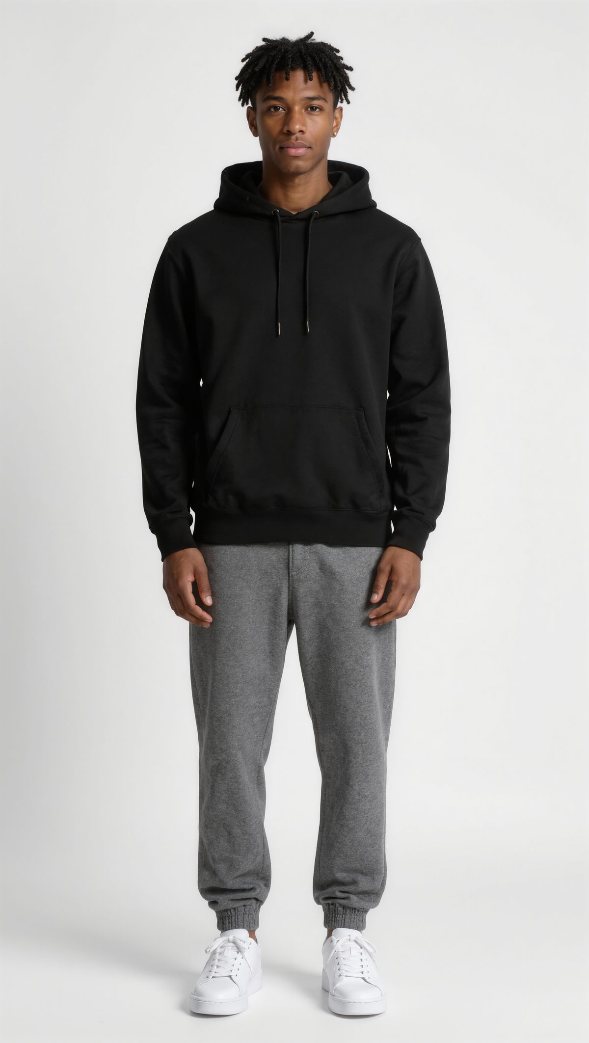 Full-length portrait of a young man with short twists in a black hoodie, gray joggers, and white sneakers