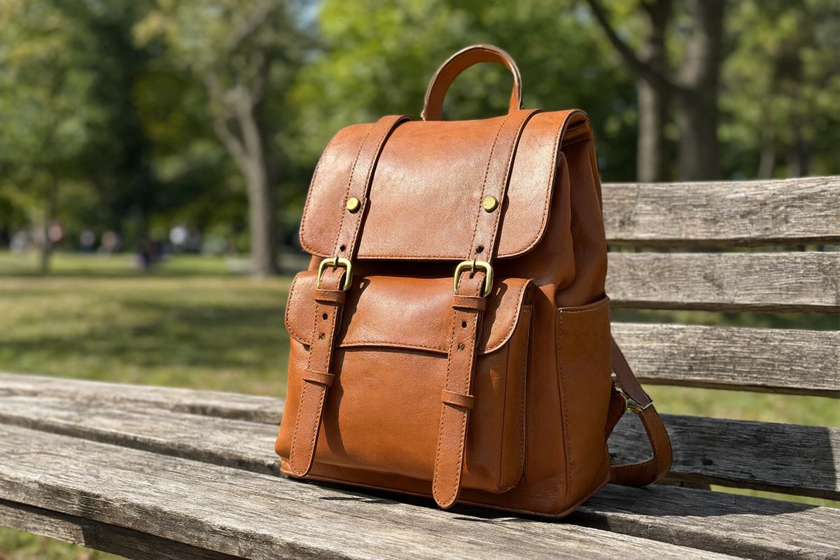 A tan leather backpack with brass buckles standing on a wooden bench in a sunlit park