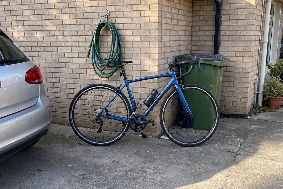 A blue road bike leaning against a light brick wall on a concrete driveway, a green wheelie bin behind the rear wheel, a coiled garden hose on the wall, and the rear of a silver car at the left edge