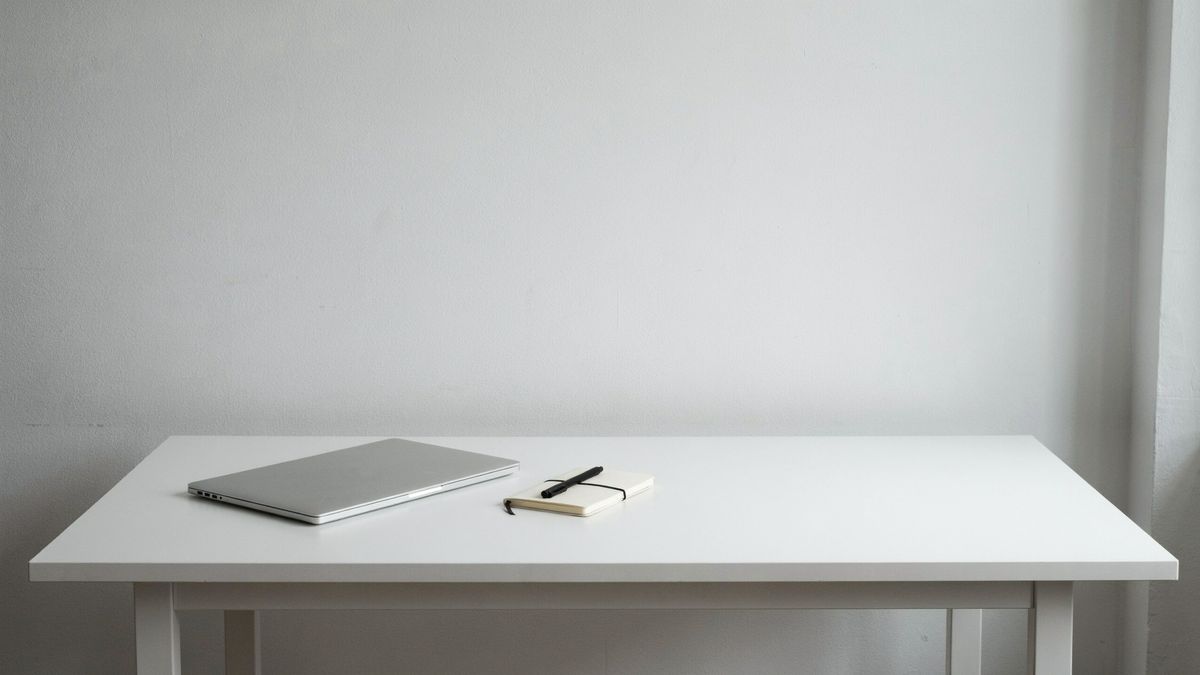 A white desk against a light gray wall, with a laptop and a notebook on the left and a tall masked area just right of the notebook