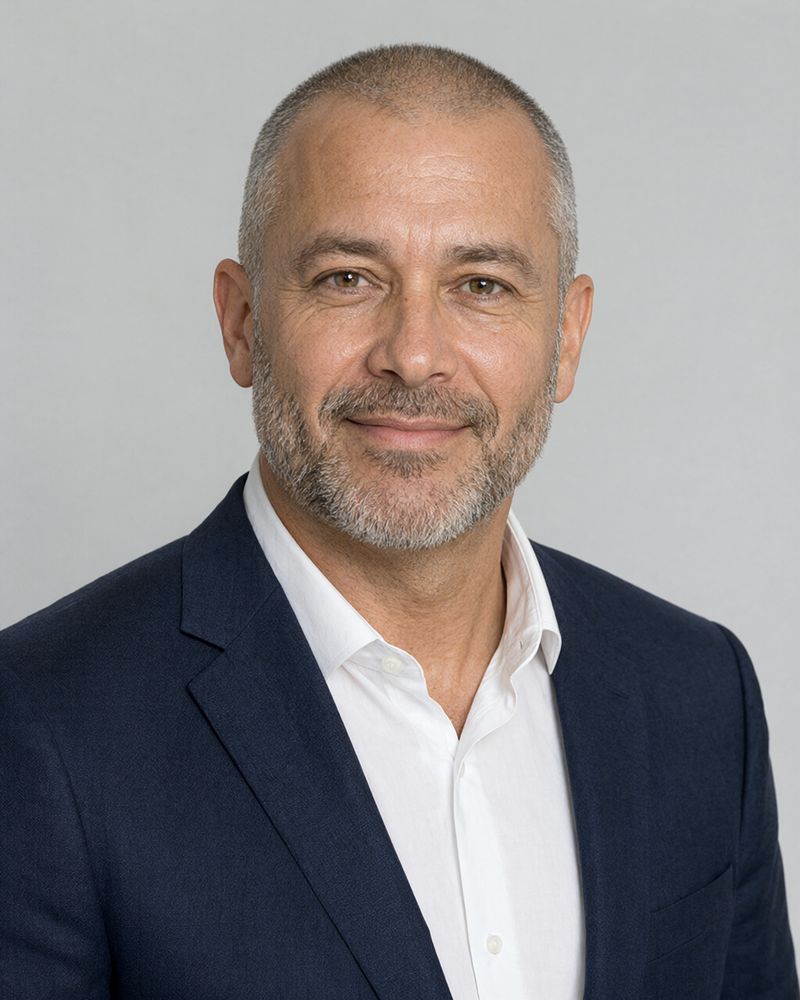 A man in his fifties with close-cropped gray hair and a gray beard in a navy blazer and white shirt