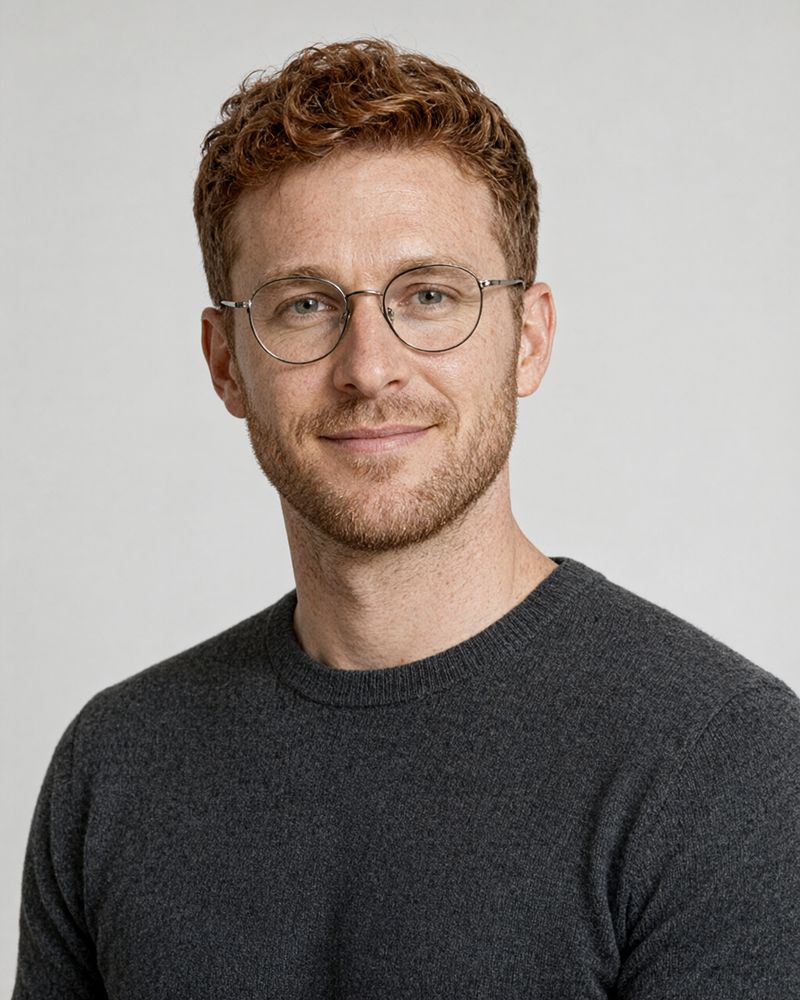 A man in his late twenties with short curly red hair and round wire glasses in a charcoal sweater