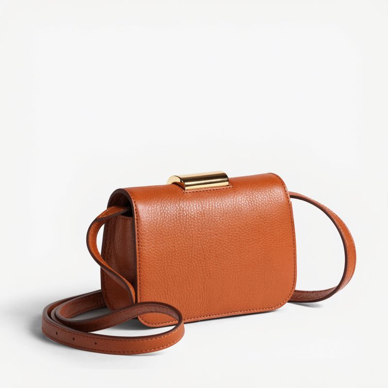 A caramel-brown pebbled leather crossbody handbag with a slim gold buckle on a plain off-white studio backdrop