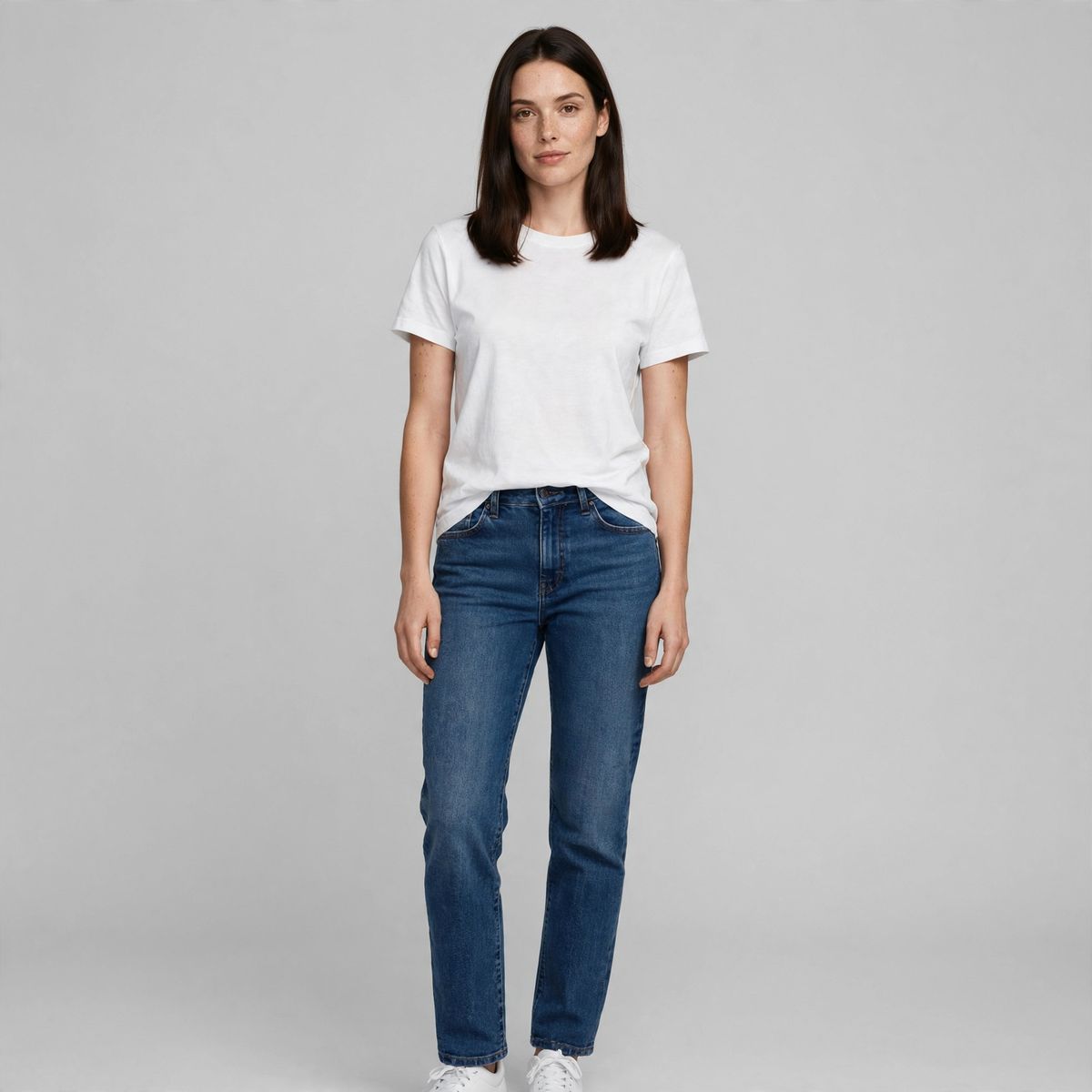 A full-length studio photo of a woman in a white t-shirt and indigo jeans standing on a plain light grey backdrop