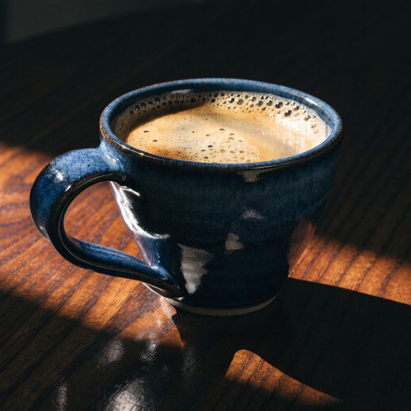 Dramatic chiaroscuro product photograph of a hand-thrown ceramic coffee mug in matte navy glaze on a dark walnut surface, single warm directional light, dark chocolate background falling to black