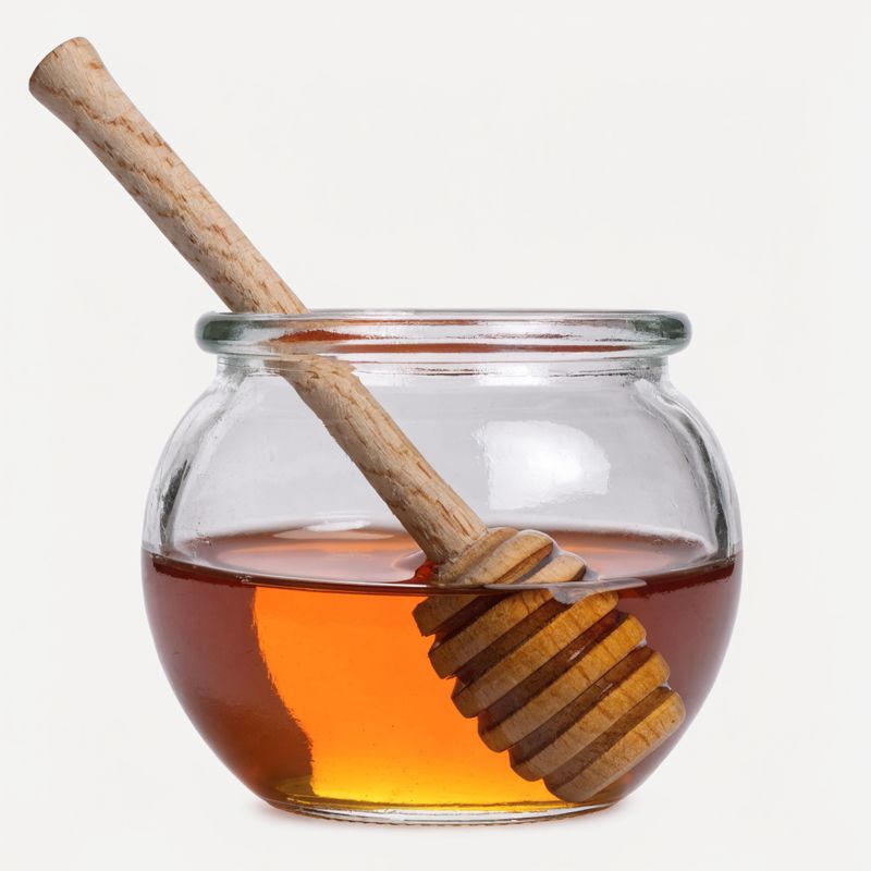 A small round glass jar half-filled with amber honey and a birch wood honey dipper resting inside, on a plain off-white studio backdrop