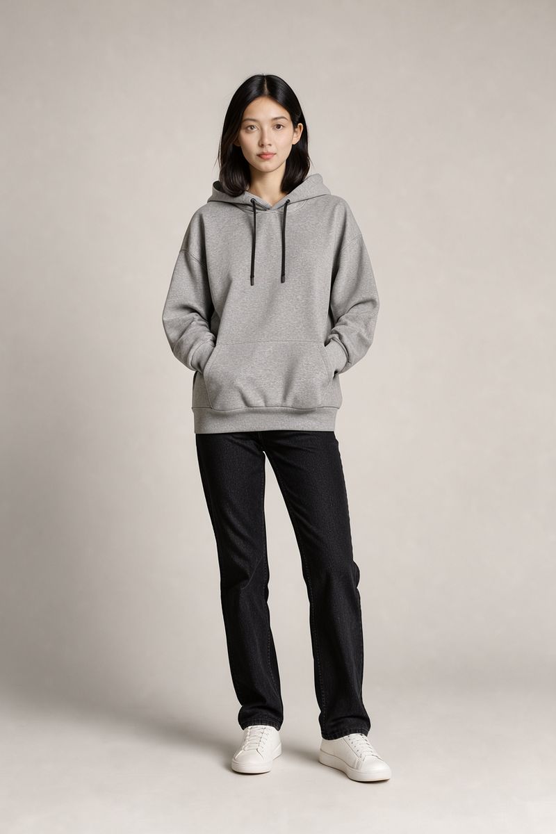 A woman with shoulder-length black hair in a heather gray oversized hoodie, black jeans and white sneakers standing on a light gray studio backdrop