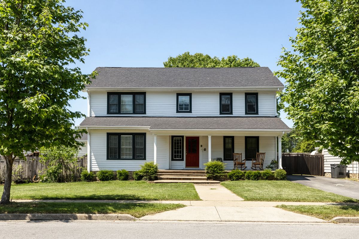 A white two-story house with a red front door, a covered porch, a green lawn, and leafy maple trees under a blue sky
