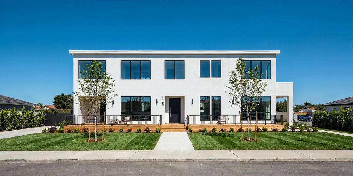A wide photograph of a modern two-story house with white stucco walls, black-framed windows and a wooden deck behind a green lawn
