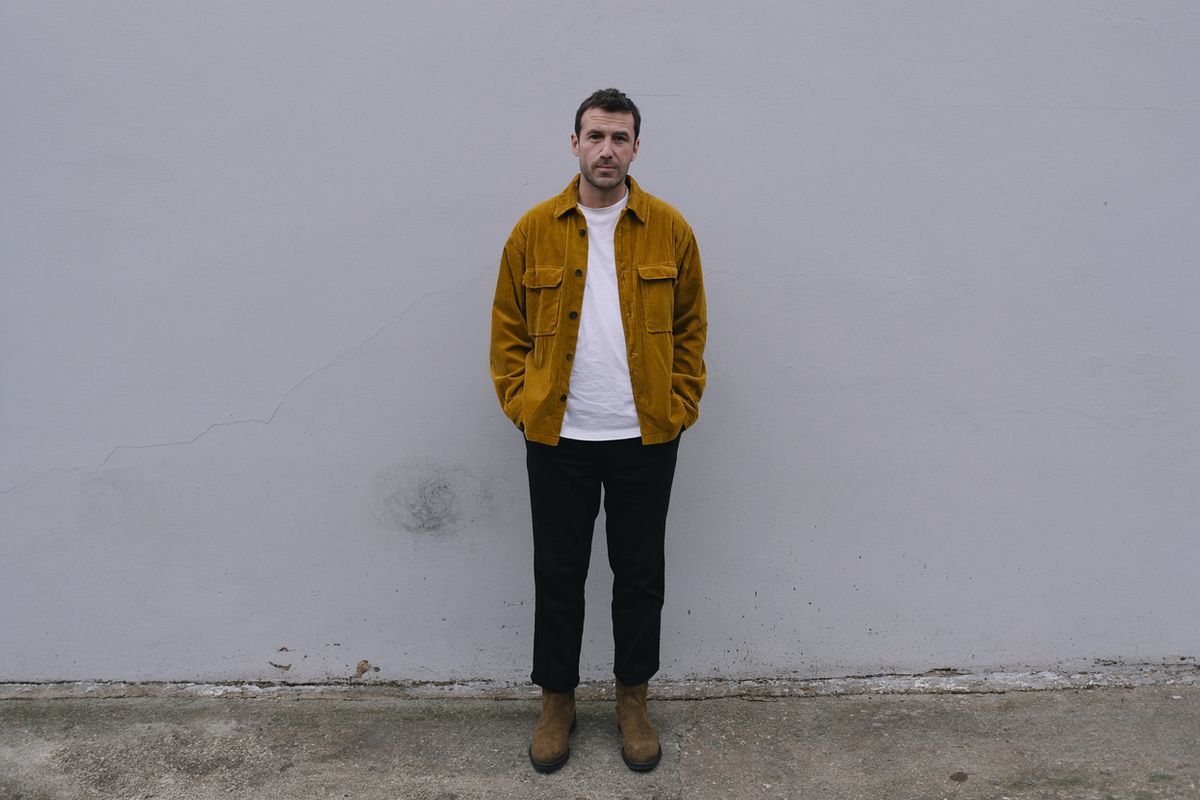 A man in a mustard yellow corduroy overshirt and black trousers standing against a plaster wall