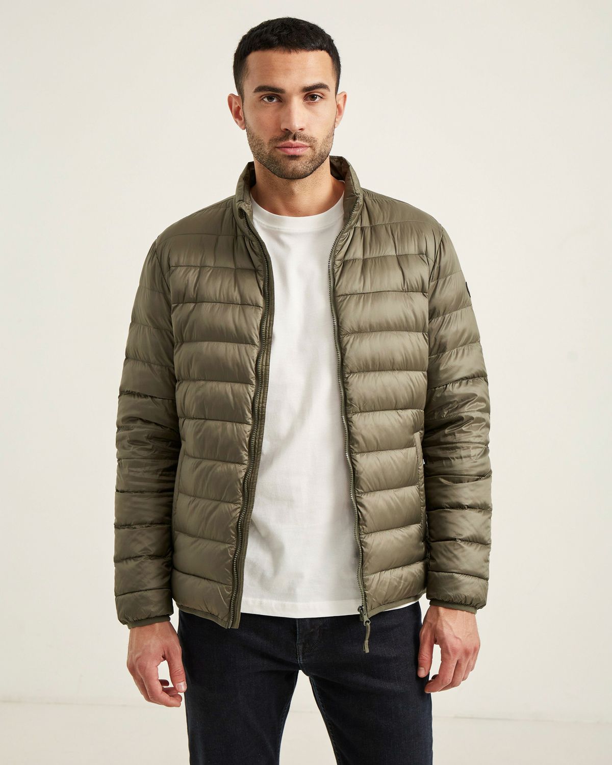 A man in an olive green quilted puffer jacket over a white t-shirt and dark jeans, standing against a warm white studio wall