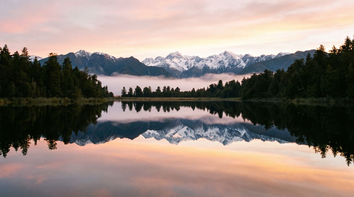 A still photograph of a mist-covered alpine lake at dawn, the still water mirroring snow-capped peaks and dark pines along the shore