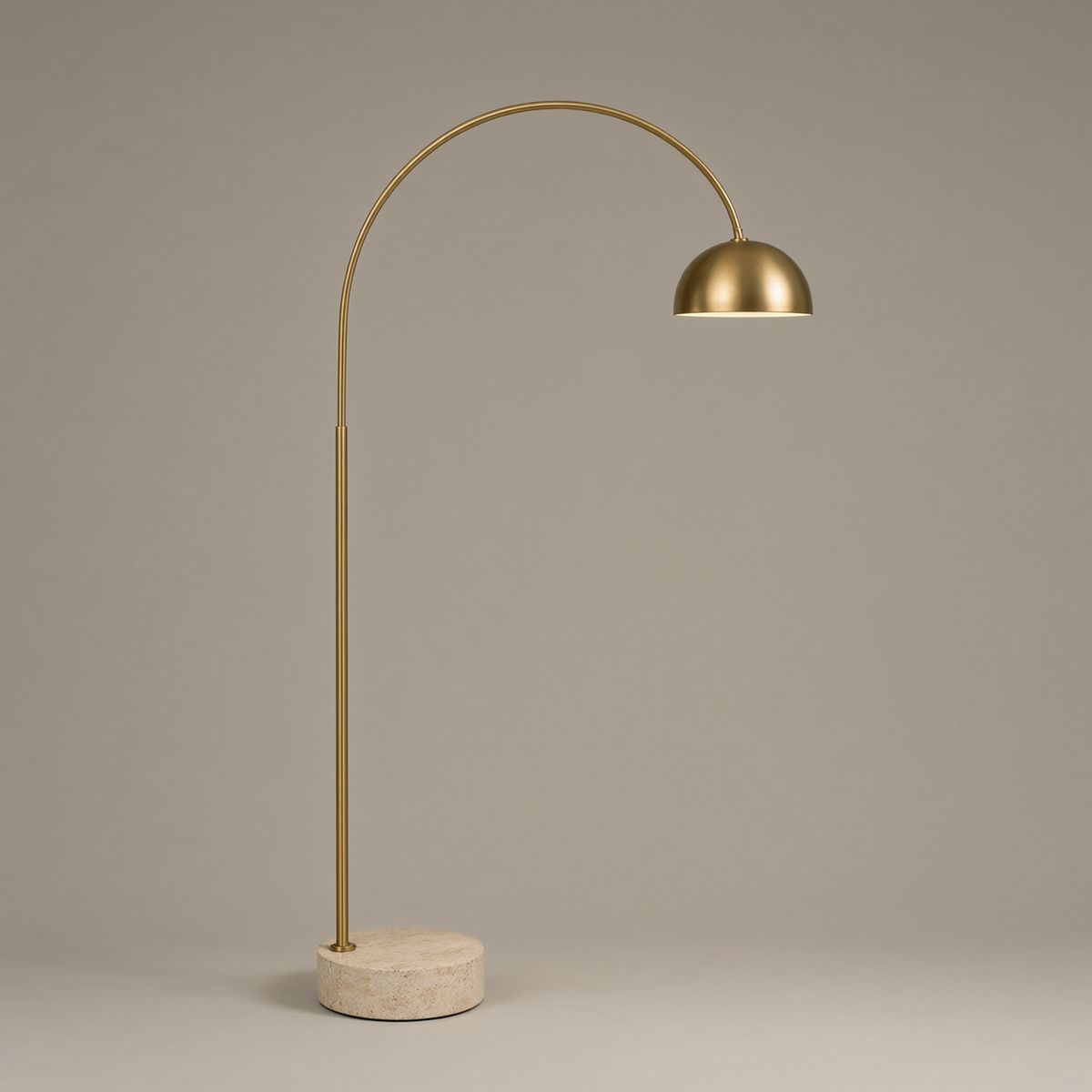 A brass arc floor lamp with a dome shade and a round travertine base on a plain warm grey backdrop
