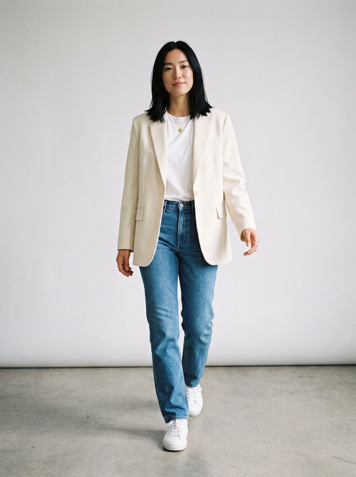 A woman with shoulder-length black hair walking toward the camera in a cream blazer, white T-shirt, blue jeans, and white sneakers against a light gray studio backdrop