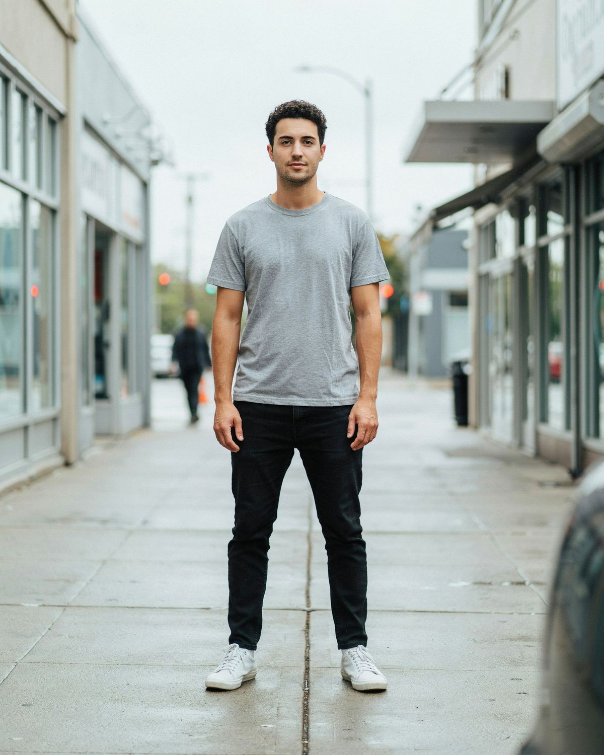 A man in a gray t-shirt, black jeans and white sneakers standing on a city sidewalk