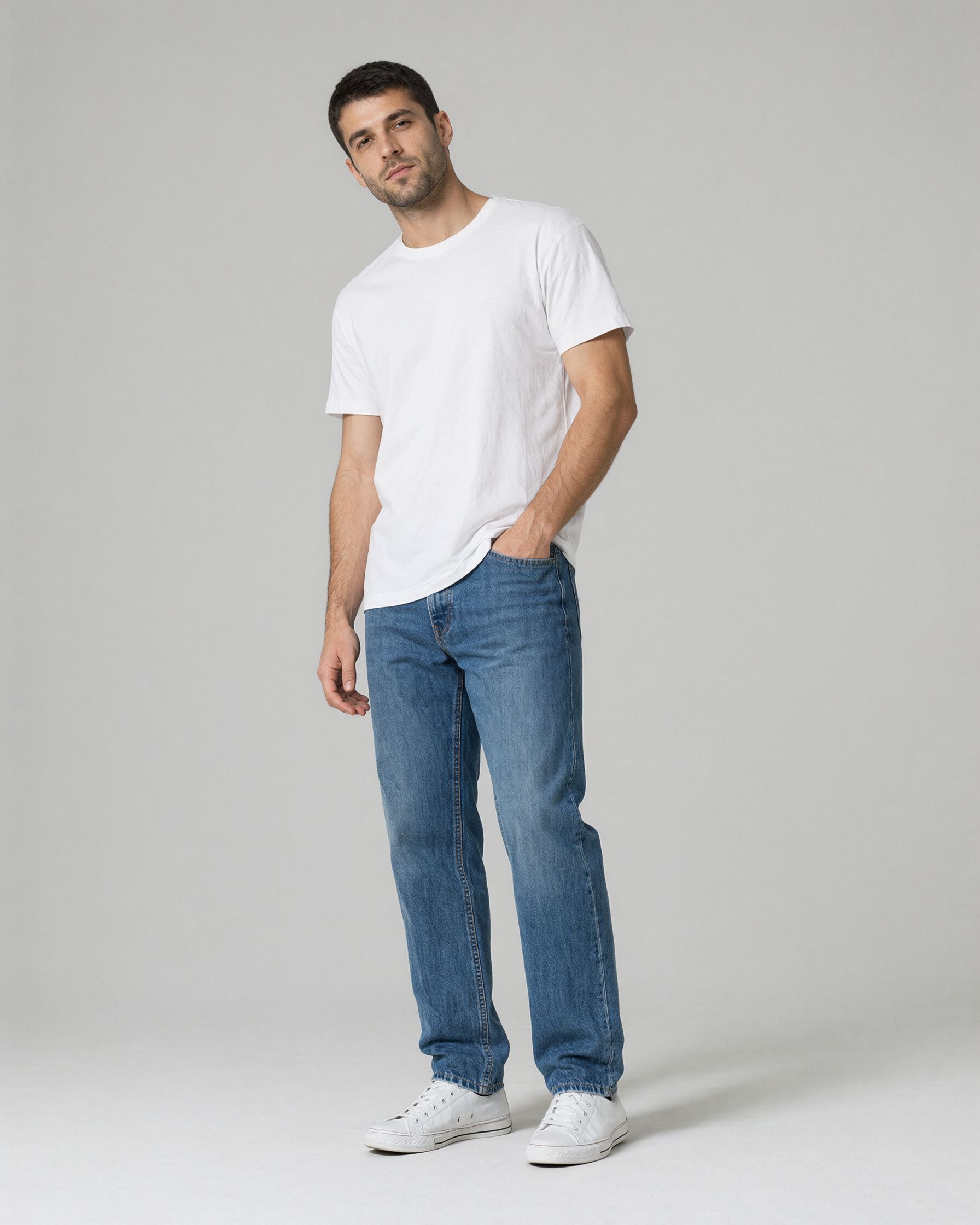 A man with short dark hair and a trimmed beard in a white T-shirt, blue jeans, and white sneakers, standing in a relaxed three-quarter stance with one hand in his pocket against a light gray backdrop