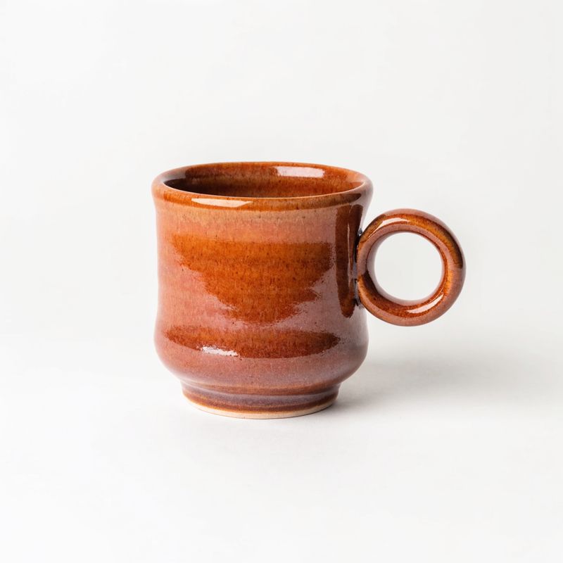 A hand-thrown stoneware coffee mug in a warm terracotta glaze with a small round handle on a plain off-white studio backdrop