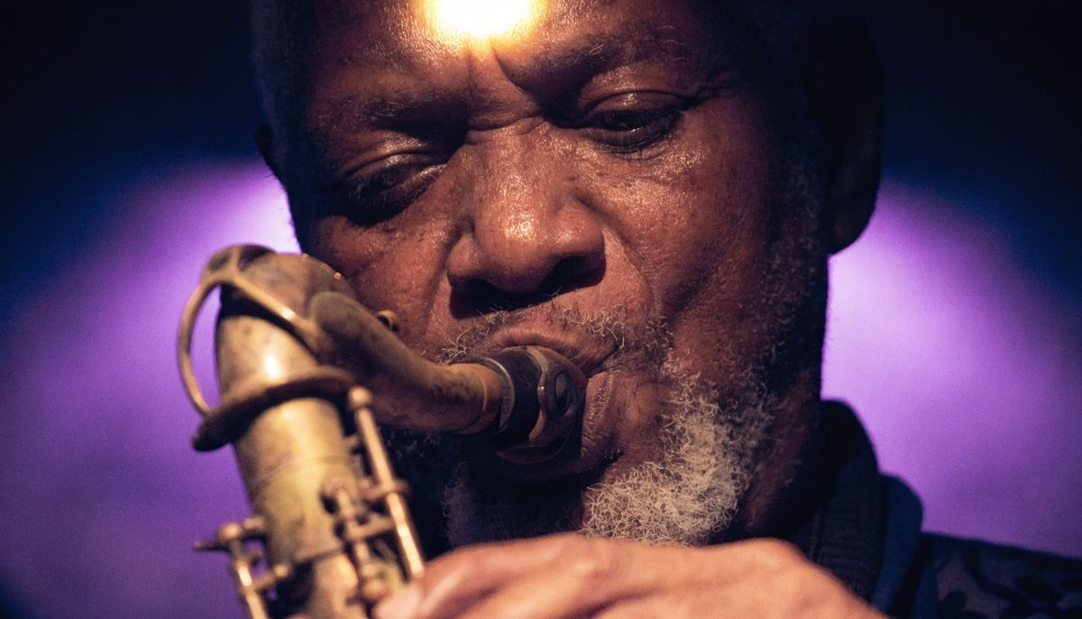 Close portrait of an elderly Black jazz saxophonist playing a tenor saxophone in deep stage lighting