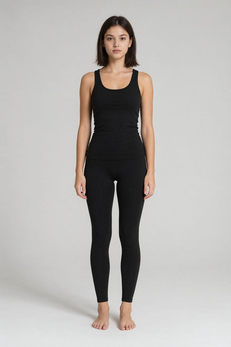 A woman with shoulder-length dark brown hair in a black tank top and black leggings standing barefoot against a light gray backdrop