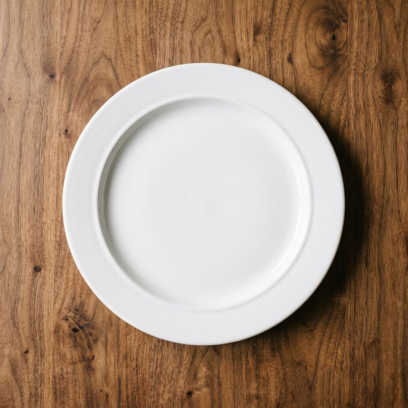 An empty pristine white ceramic plate on a warm walnut table, overhead angle, soft warm kitchen light