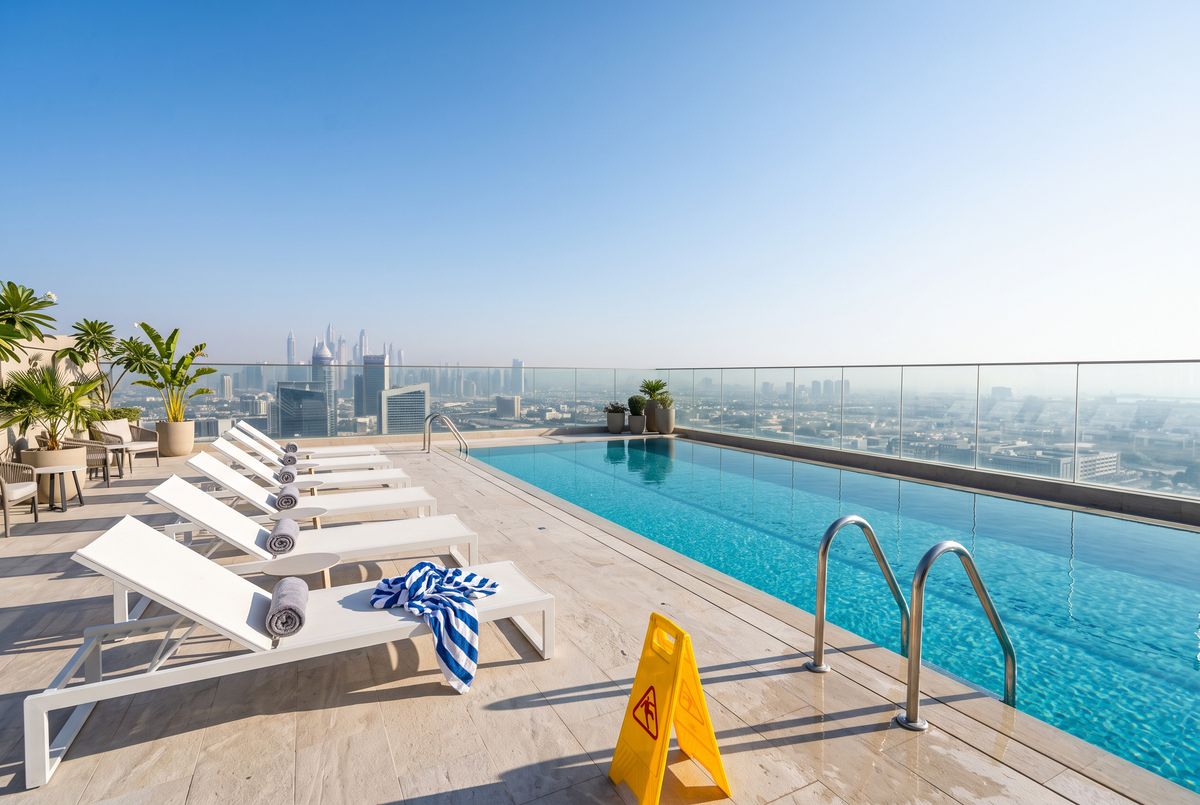 A rooftop pool deck with a turquoise pool on the right, a row of white loungers on the left, a city skyline behind a glass balustrade, a yellow wet-floor sign by the pool steps, and a crumpled striped towel on the nearest lounger