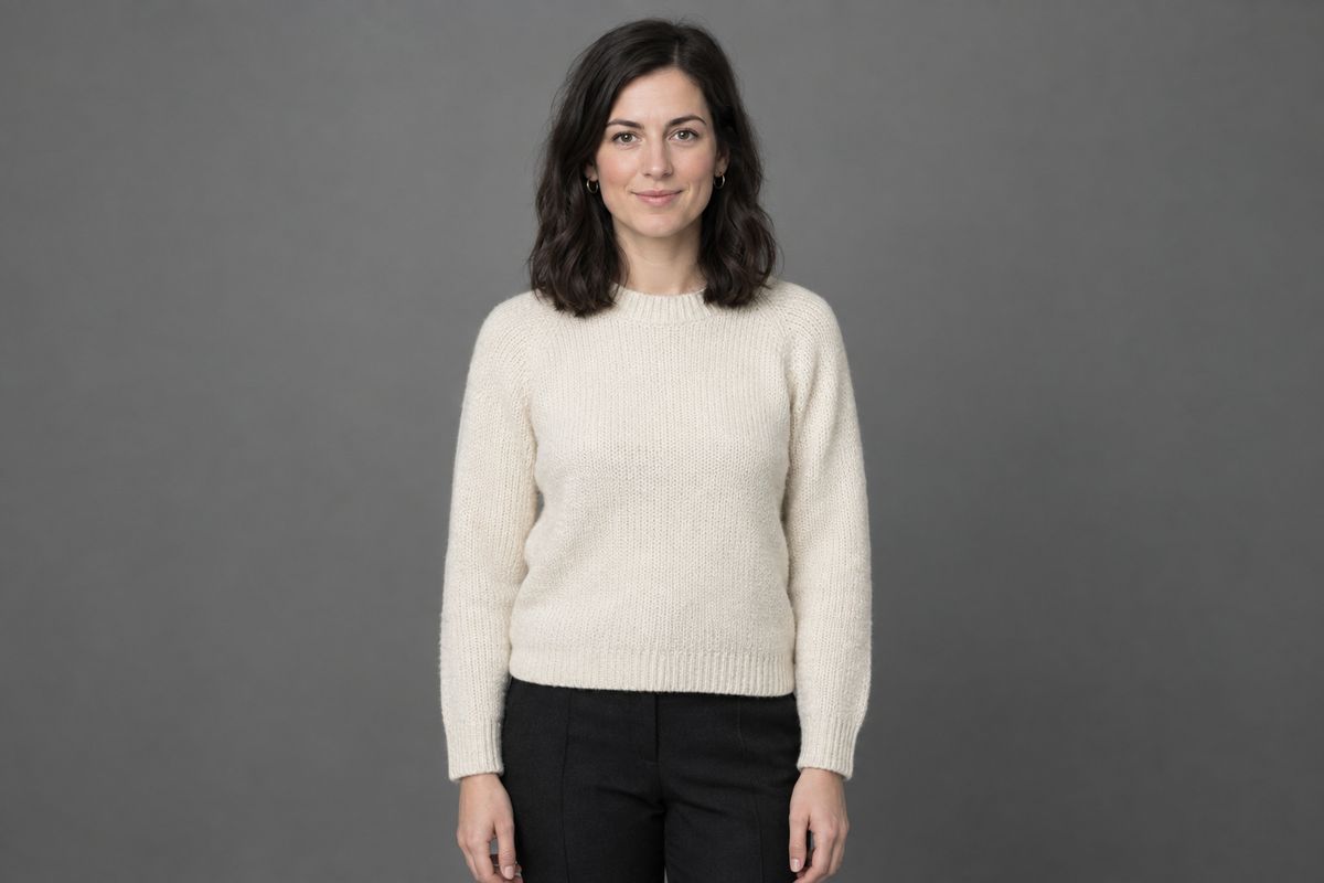 A woman standing against a plain grey studio wall in a cream knitted jumper and dark trousers, facing the camera