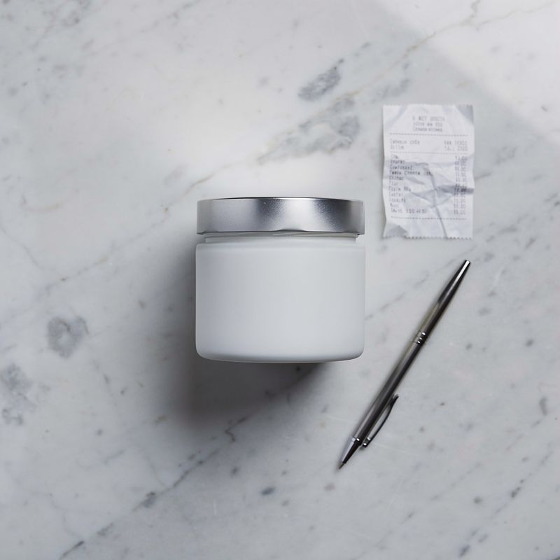 Top-down flat-lay of a white skincare jar on marble with a receipt and pen nearby