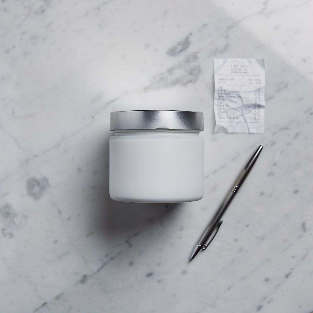 Top-down flat-lay of a white skincare jar on marble with a receipt and pen nearby