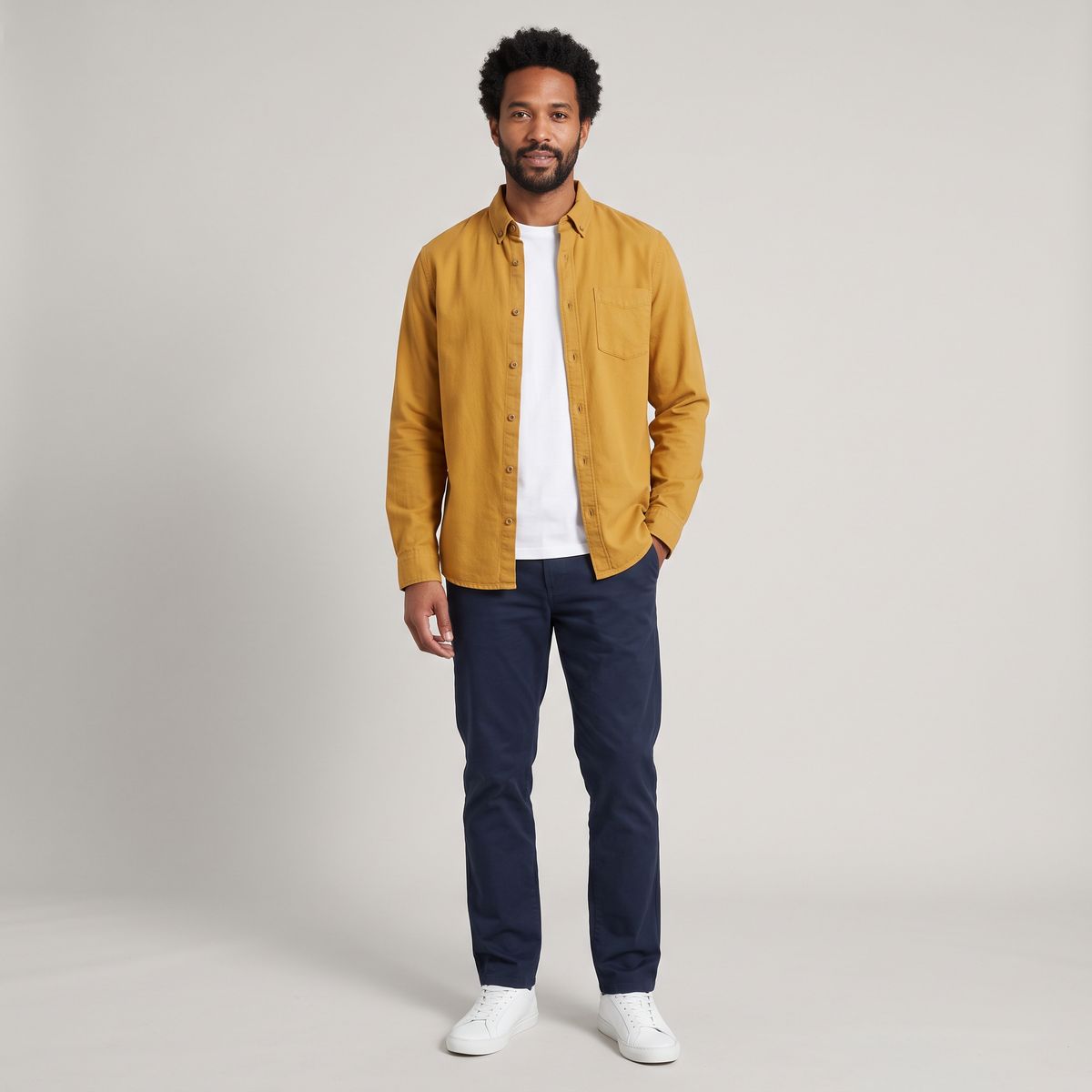 A man with short curly black hair and a trimmed beard standing in a mustard yellow overshirt, white T-shirt, navy chinos, and white sneakers against a light gray backdrop