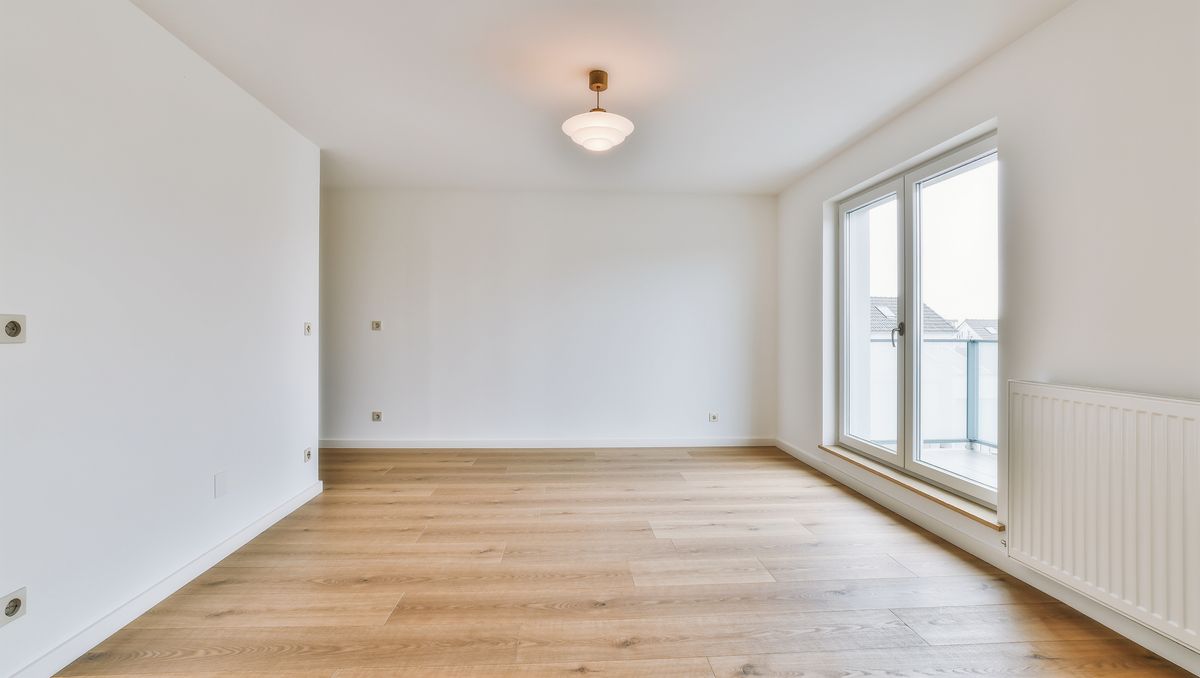 An empty renovated living room with white walls, a light oak floor, and a large window