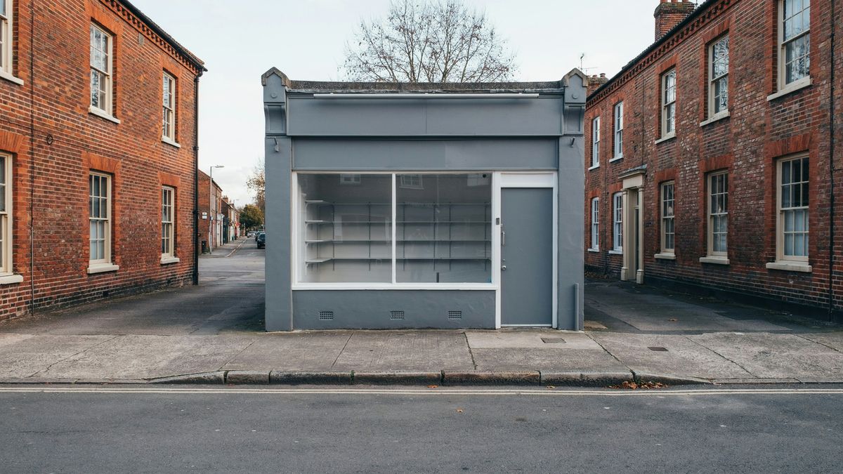 A small shop front with a dark gray facade, a blank sign board, an empty display window and a gray metal door, between two red brick buildings