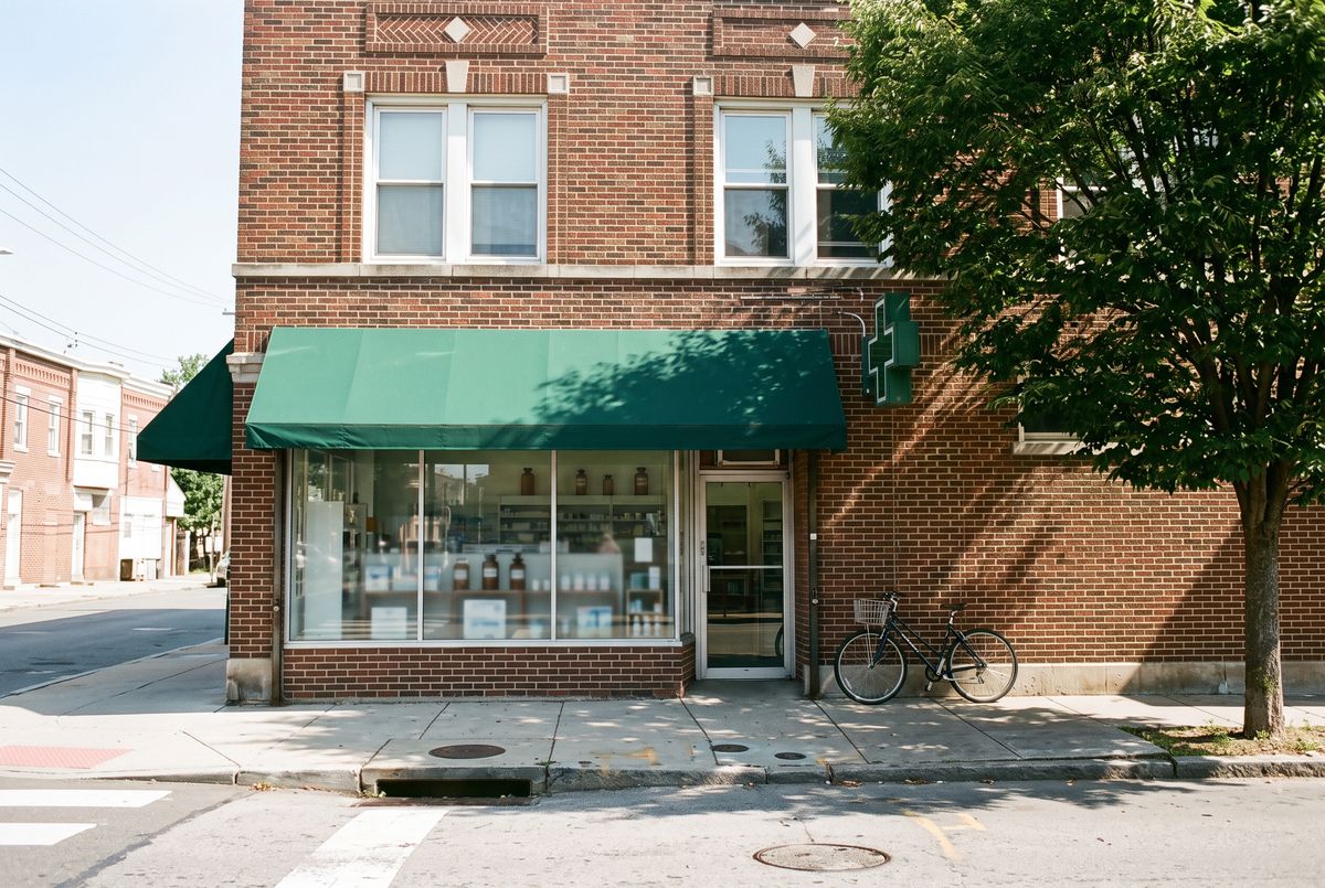 A photograph of a two-story brick corner pharmacy with a large display window, a green awning, a green cross sign, a bicycle by the door, and a street tree