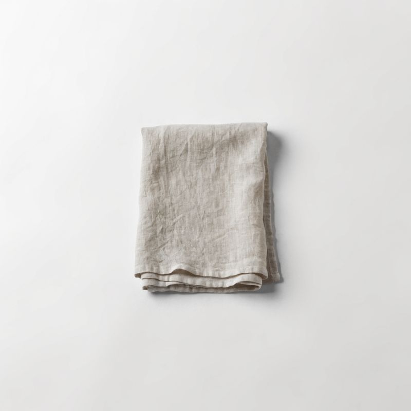 A folded natural undyed linen tea towel with visible weave texture, laid flat on a plain off-white studio backdrop