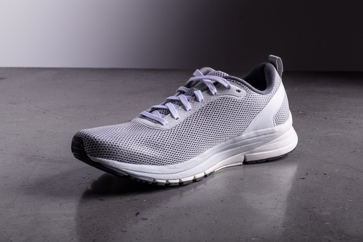 A gray and white mesh running shoe on a dark concrete floor, lit from the side against a shadowed backdrop
