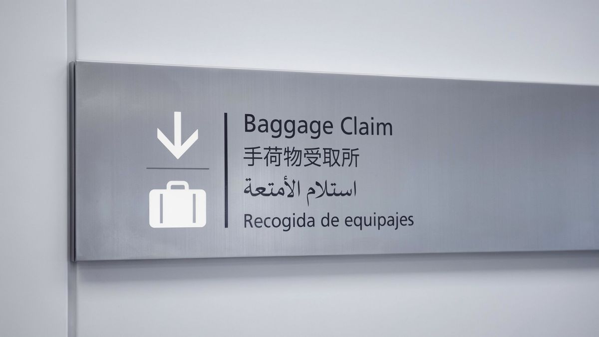 A brushed-aluminium airport wayfinding sign reading 'Baggage Claim' in English, 手荷物受取所 in Japanese, استلام الأمتعة in Arabic, and 'Recogida de equipajes' in Spanish, alongside a white suitcase pictogram and downward arrow