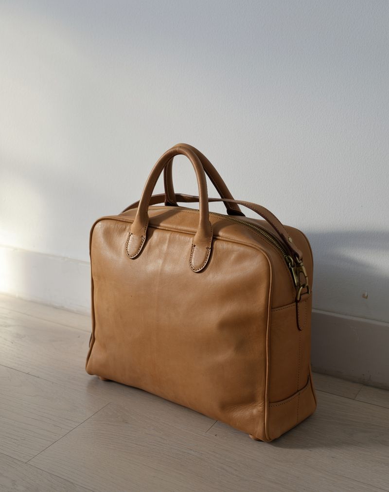 A tan leather weekender bag standing on a bleached oak floor beside a white wall, lit from the left